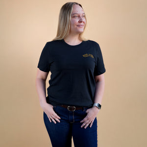Thumbnail of Woman wearing a black t-shirt with a yellow logo against a beige background