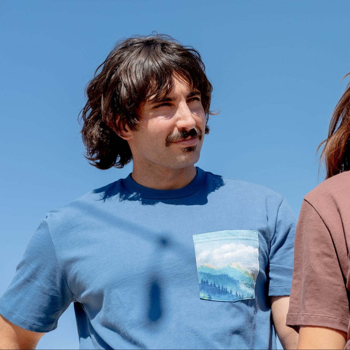 Two people wearing t-shirts with a scenic design against a clear blue sky.