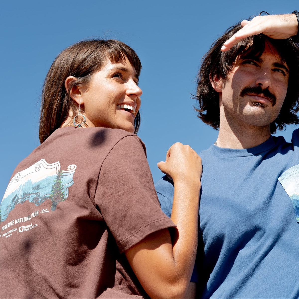 Two people wearing t-shirts with nature-themed designs against a clear blue sky.