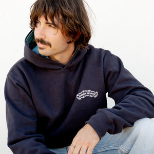Thumbnail of Man wearing a navy hoodie with a logo, sitting on a concrete surface.