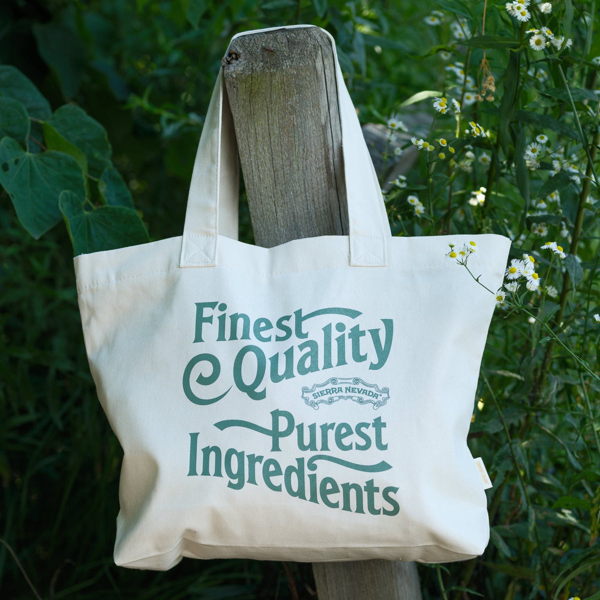 An off-white canvas tote bag with green printed text reading 'Finest Quality Purest Ingredients' hanging from a wooden post in a garden setting.