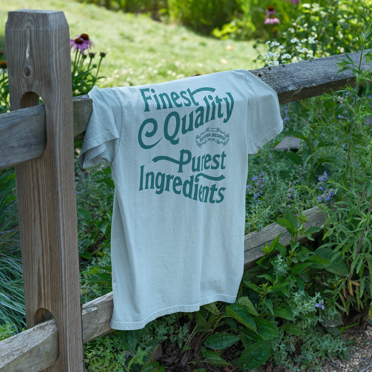A light gray t-shirt with a green graphic print reading 'Finest Quality Purest Ingredients' draped over a wooden fence in a lush green garden setting.