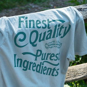 Thumbnail of A light blue-gray t-shirt draped over a rustic wooden fence outdoors, featuring a dark teal graphic print that reads 'Finest Quality Purest Ingredients' with a 'Sierra Nevada' logo.