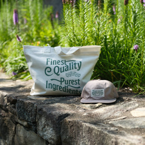 Thumbnail of A cream canvas tote bag with green text reading 'Finest Quality Purest Ingredients' sits on a stone wall next to a beige trucker cap, with a lush green garden in the background.