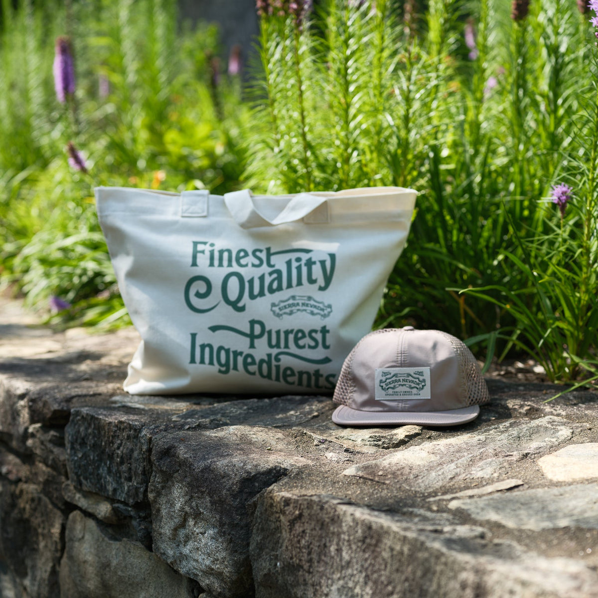 A cream canvas tote bag with green text reading 'Finest Quality Purest Ingredients' sits on a stone wall next to a beige trucker cap, with a lush green garden in the background.