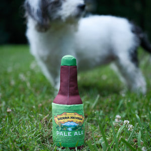 Thumbnail of A dog toy shaped like a beer bottle with a green cap and a green label reading 'Sierra Nevada Pale Ale', placed on grass with a small dog blurred in the background.