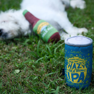 Thumbnail of A blue novelty can-shaped dog toy with yellow and white graphics reading 'HAZY LITTLE THING IPA' sits on green grass, with a white dog lying nearby in the background.