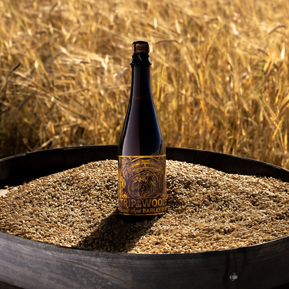 A dark glass bottle with a gold and black label reading 'Barrel-Aged Barleywine' stands on a barrel filled with grains, set against a blurred golden wheat field.