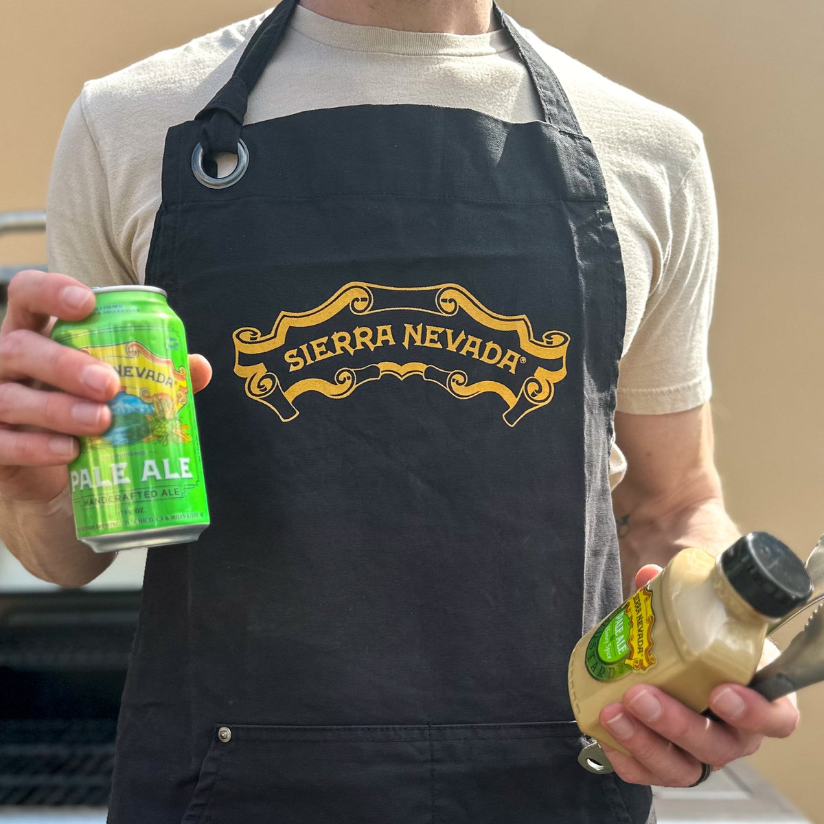 Person wearing a Sierra Nevada apron holding a beer can and a bottle with a spoon.