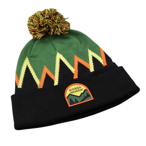 Thumbnail of A knit pom-pom beanie hat featuring a green upper section with a zigzag pattern in cream, orange, and black, a black cuff, and a Sierra Nevada mountain patch on the front.