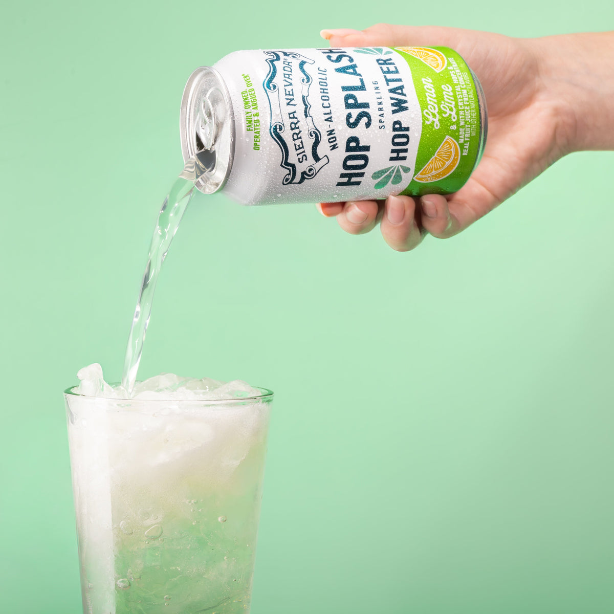 Hand pouring a can of 'Hop Splash' non-alcoholic hop water into a glass against a green background