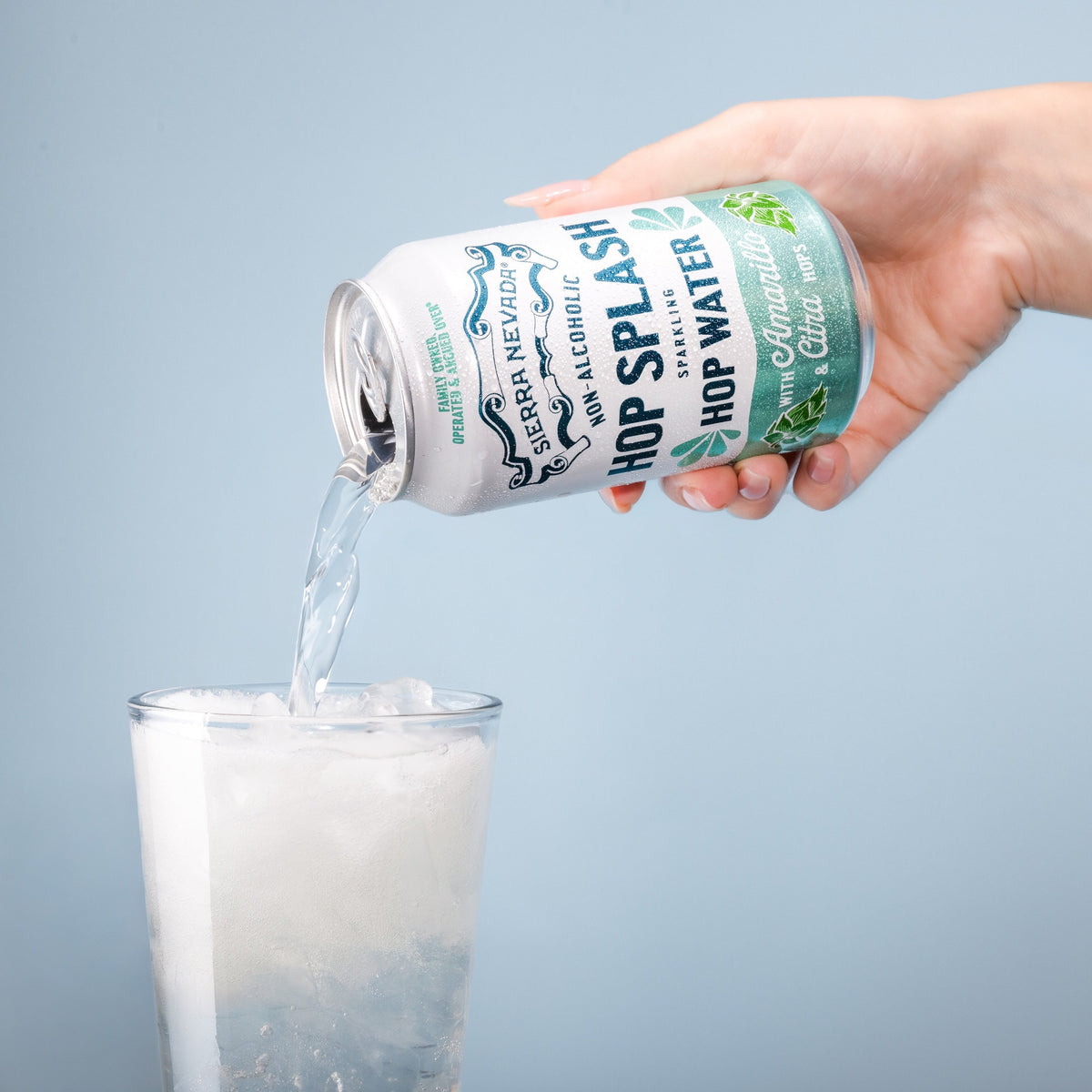 Hand pouring beer from a can labeled 'Sarver's Hop Splash' into a glass against a light blue background.