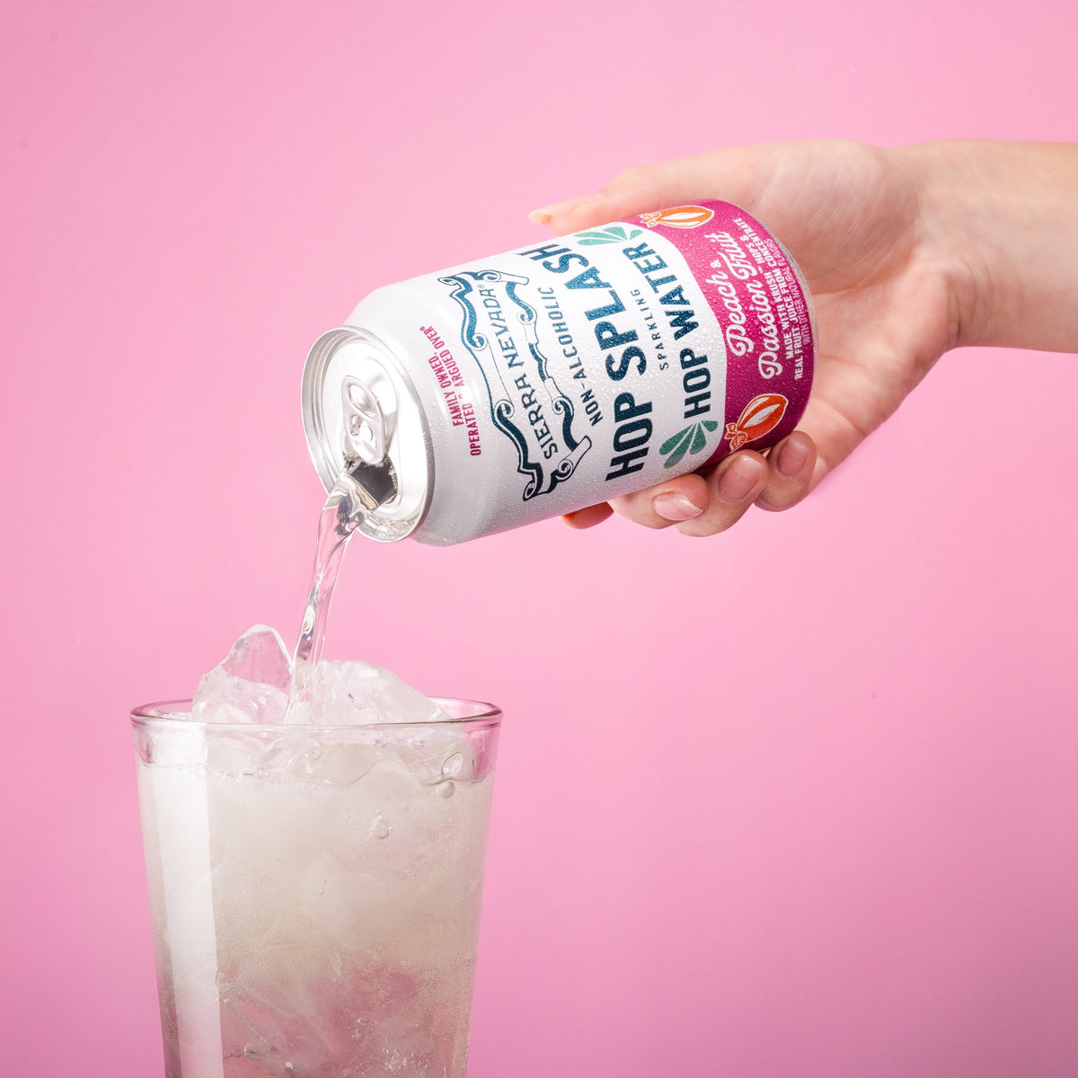 Hand pouring a can of 'Hop Splash' hot water into a glass against a pink background