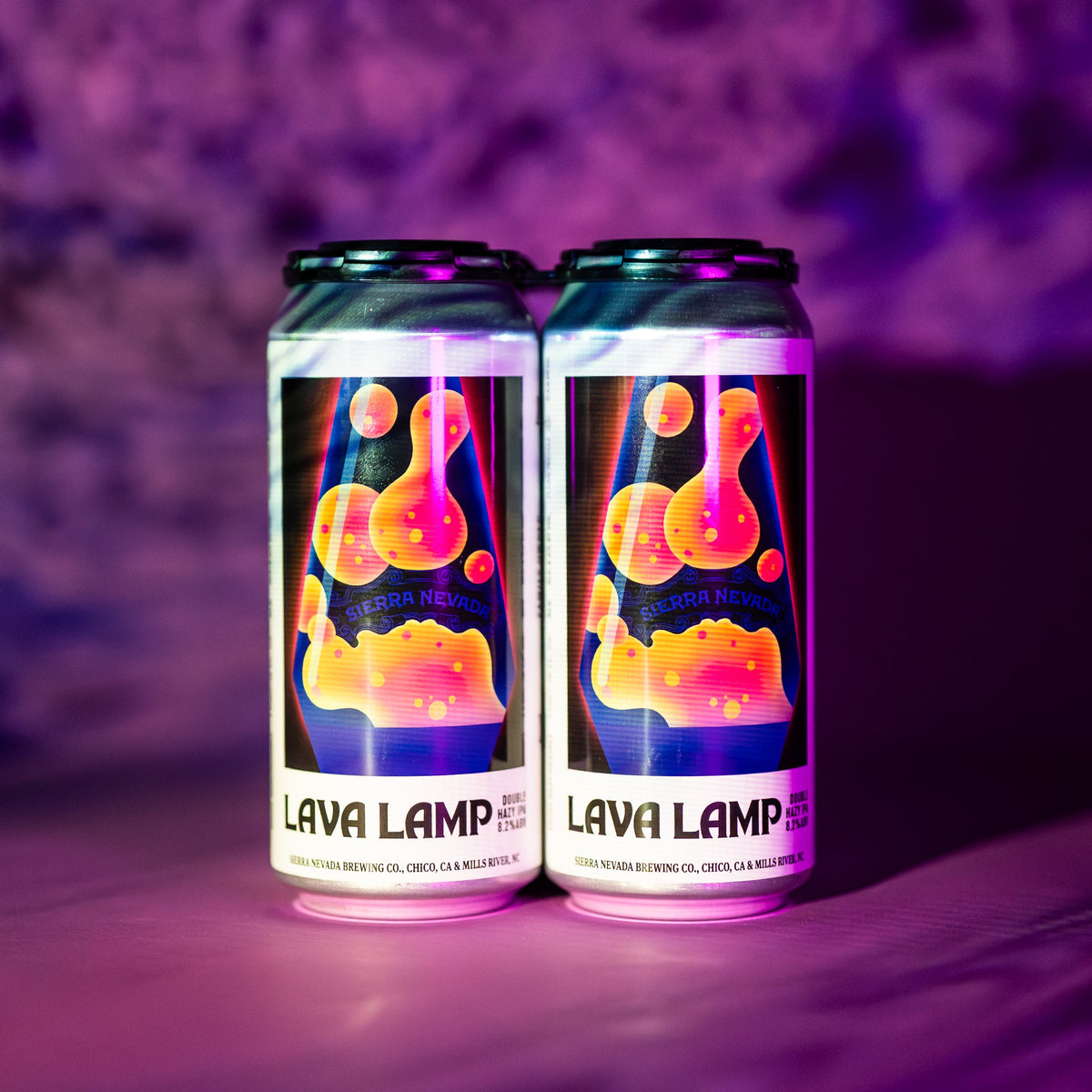 Two 16 oz aluminum cans of Lava Lamp beer on a reflective surface with a purple, smoky background. The cans feature a colorful retro lava lamp design with orange, yellow, and pink shapes on a dark background.