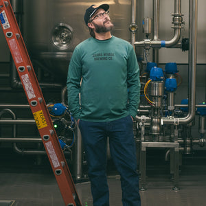 Thumbnail of Man in a teal shirt standing next to industrial brewing equipment