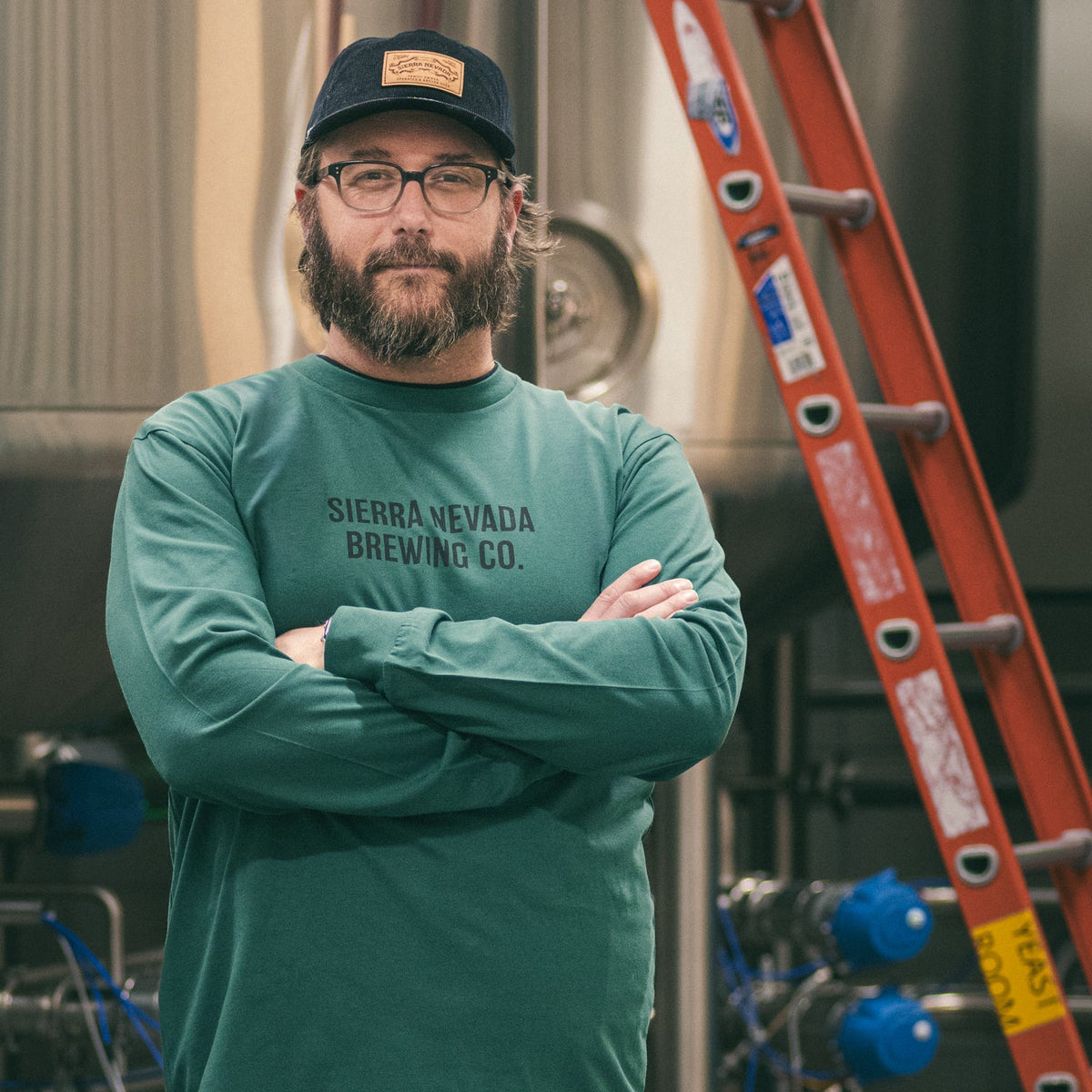 Man wearing a green Sierra Nevada Brewing Co. shirt standing in front of brewing equipment.
