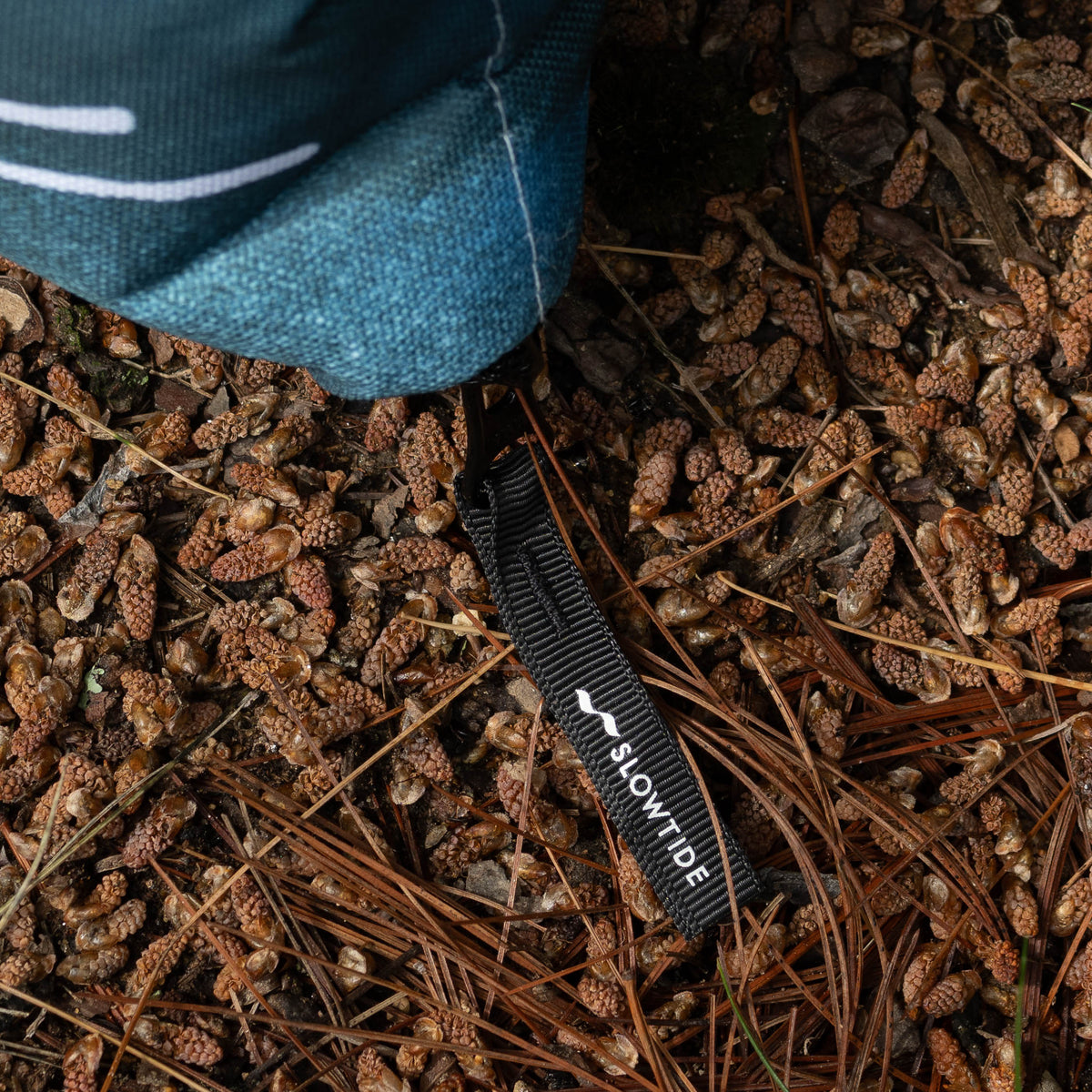 Close-up of the corner of the NPF X Sierra Nevada packable blanket with a visible brand logo on a pine cone-strewn ground.