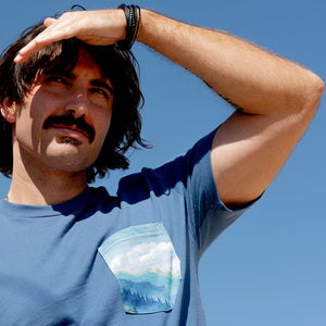 Thumbnail of Man wearing a blue t-shirt with a nature-themed pocket design against a clear blue sky.