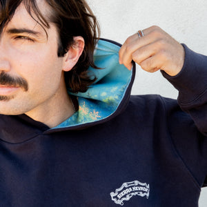 Thumbnail of Man wearing a navy blue hoodie with a visible brand logo, holding the hood up.
