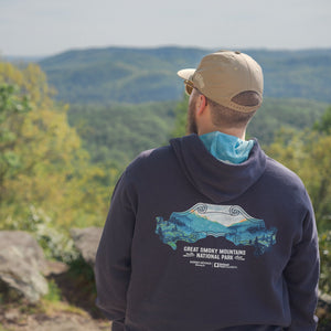 Thumbnail of Person wearing a hoodie with a mountain design, standing on a rocky outcrop with a scenic view of trees and mountains.