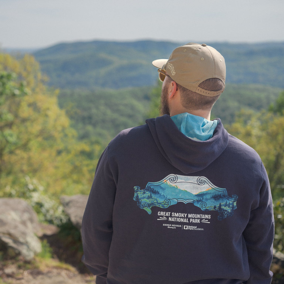 Person wearing a hoodie with a mountain design, standing on a rocky outcrop with a scenic view of trees and mountains.