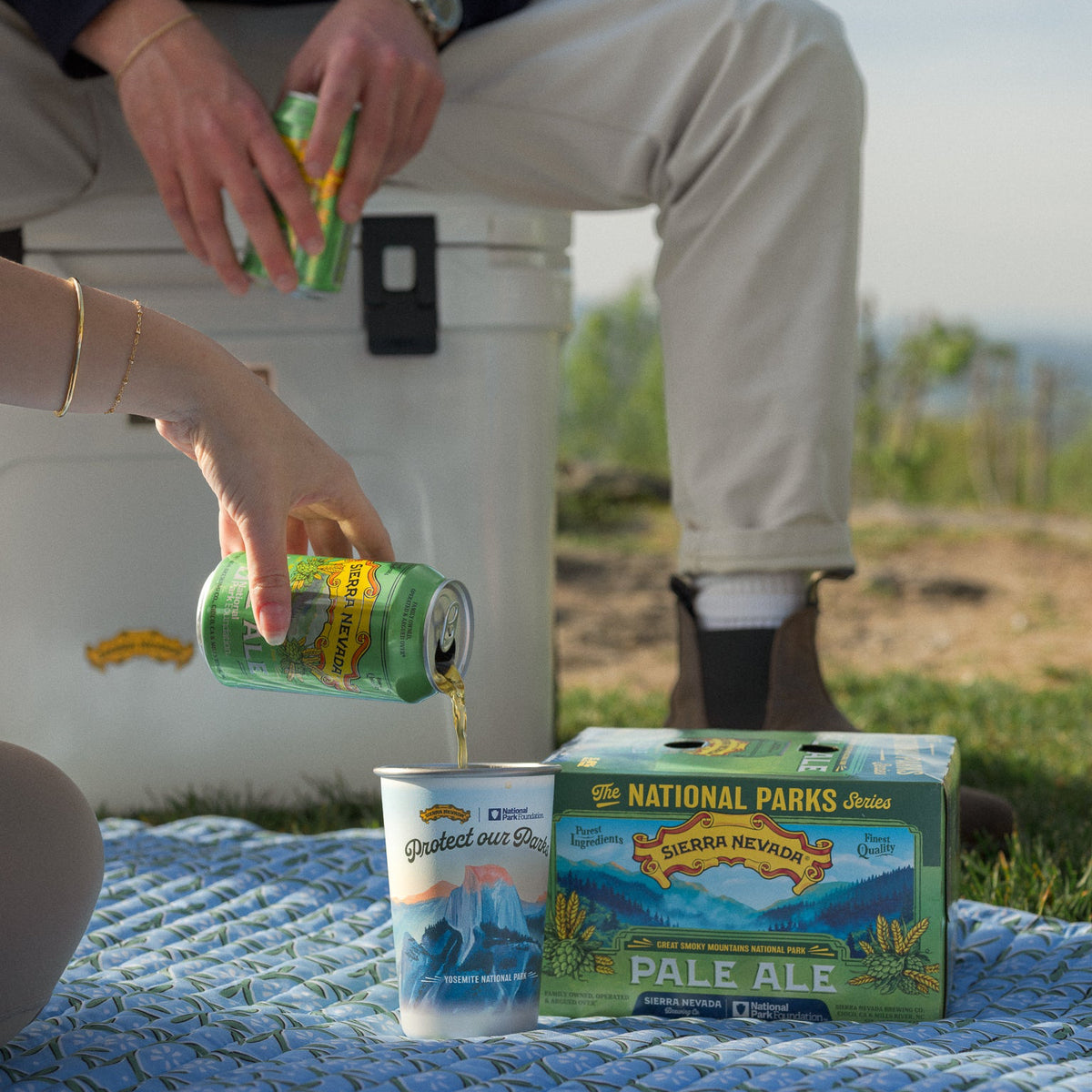 Person pouring beer from a can into a cup on a blanket with National Parks Pale Ale packaging.