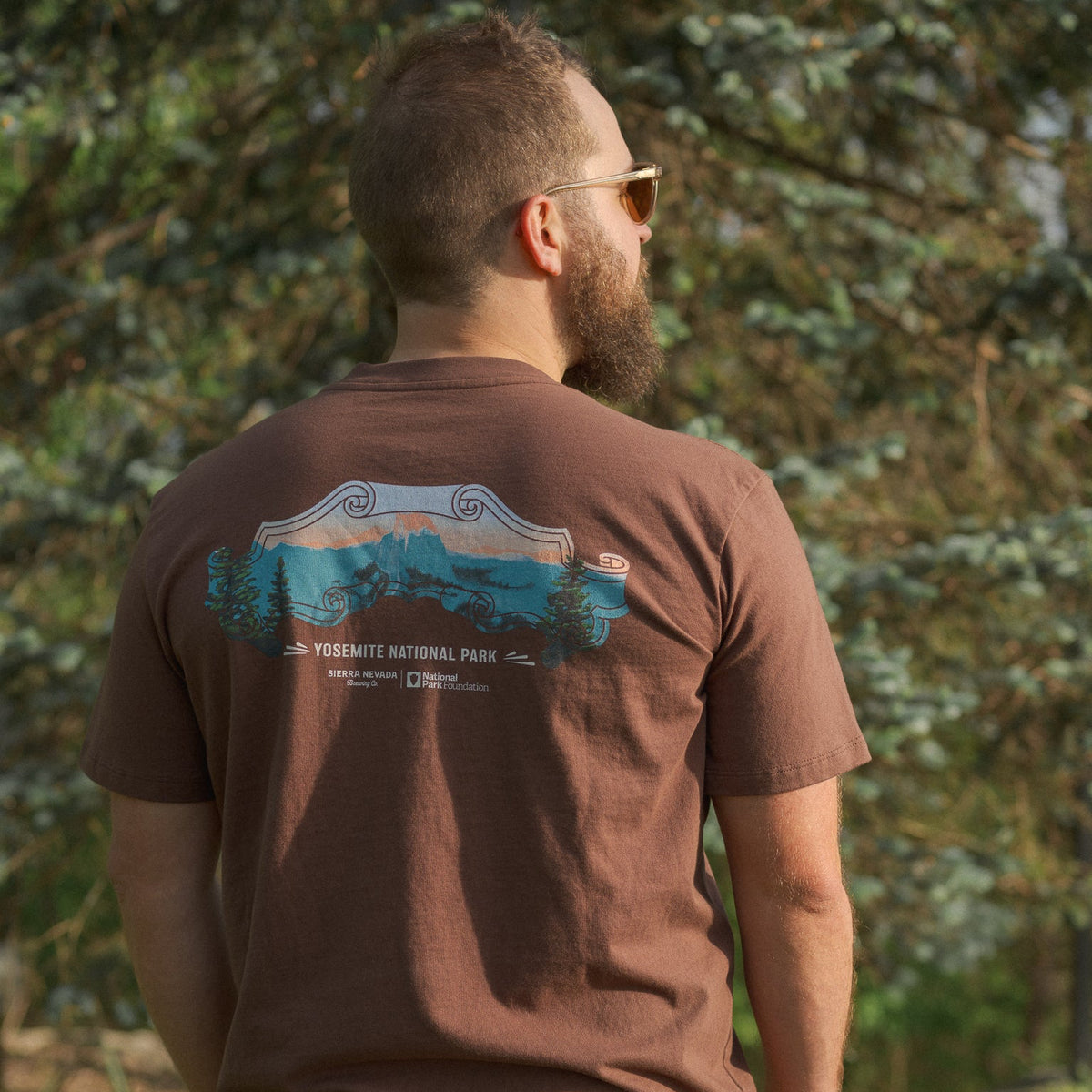 Man wearing a brown t-shirt with a nature graphic, standing in a forested area.