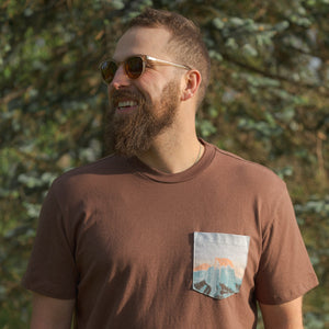 Thumbnail of Man wearing a brown t-shirt with a pocket design, standing outdoors with greenery in the background.