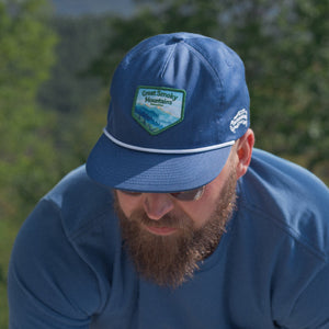 Thumbnail of Man wearing a blue cap and shirt outdoors with greenery in the background