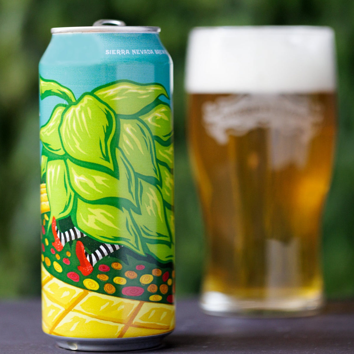 Colorful beer can with hop design next to a glass of beer on a blurred green background