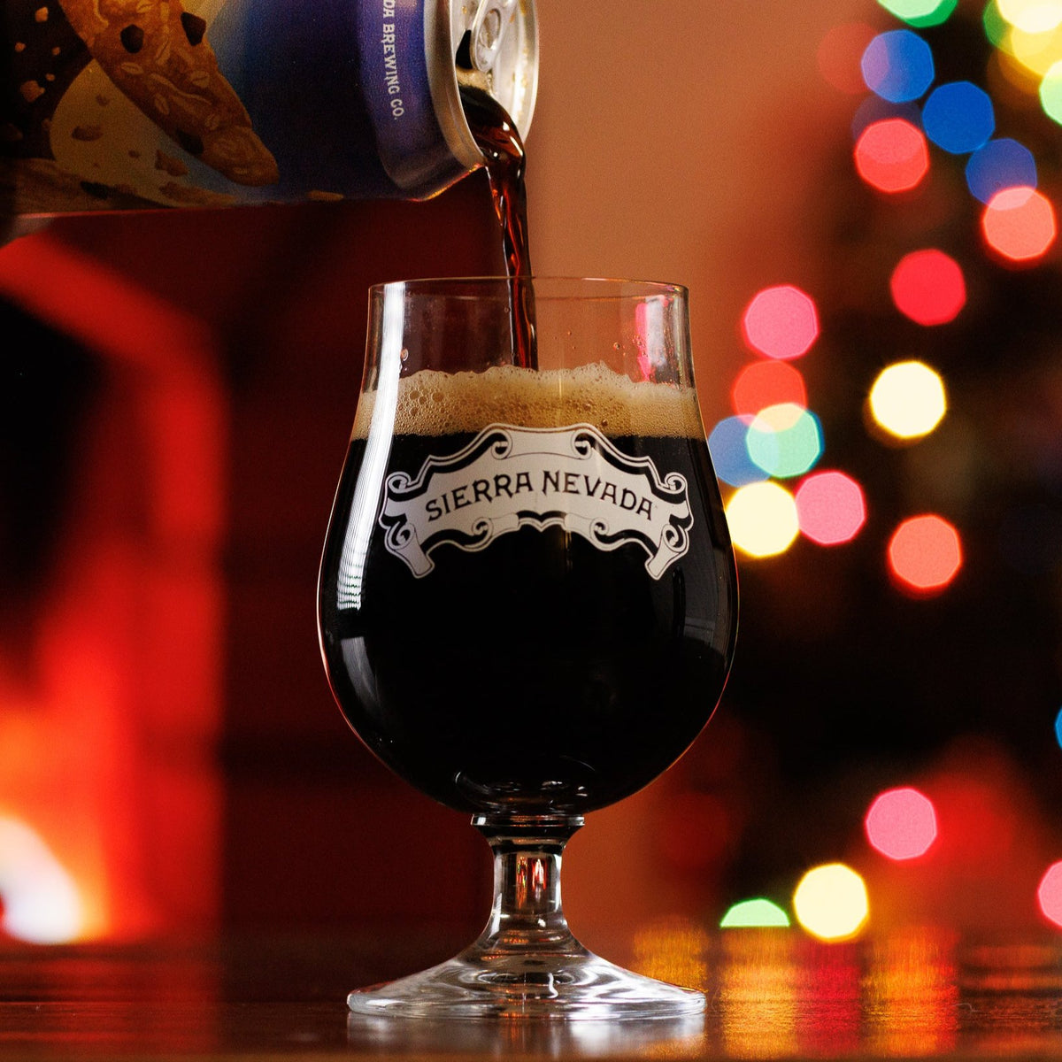 A can of Oatmeal Cookie Stout is poured into a balloon glass with festive holiday lights in the background