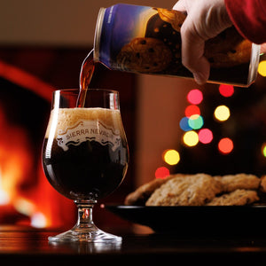 Thumbnail of A can of Oatmeal Cookie Stout is poured into a balloon glass with festive holiday lights and a plate of cookies in the background