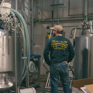 Thumbnail of Person wearing a branded shirt in a brewery setting with large metal tanks.