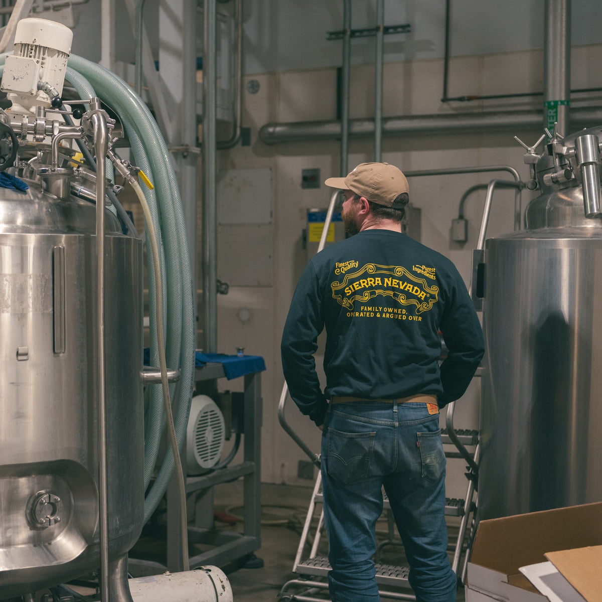 Person wearing a branded shirt in a brewery setting with large metal tanks.