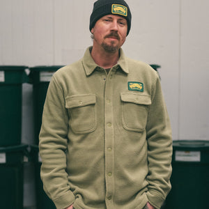 Thumbnail of Man wearing a green jacket and beanie with logos, standing in front of green bins.