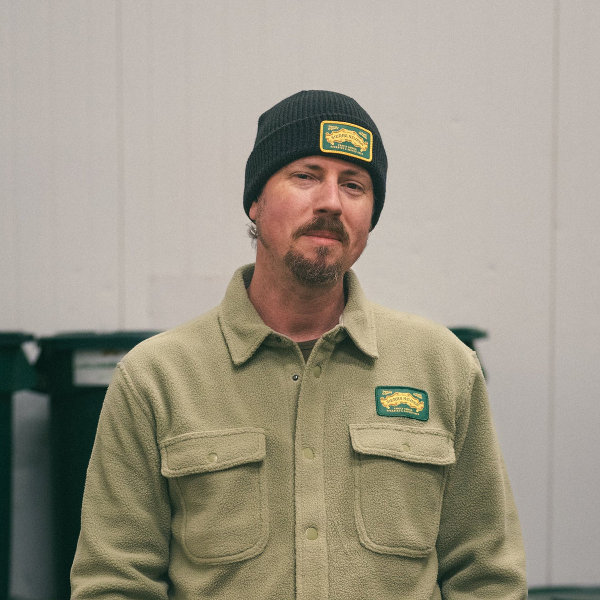 Man wearing a green jacket and beanie with logos, standing in front of green bins.