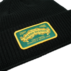 Thumbnail of Close-up of a black ribbed knit beanie with a rectangular embroidered patch reading 'Sierra Nevada' in yellow on a green background, with additional text 'Family Owned Operated & Argued Over'