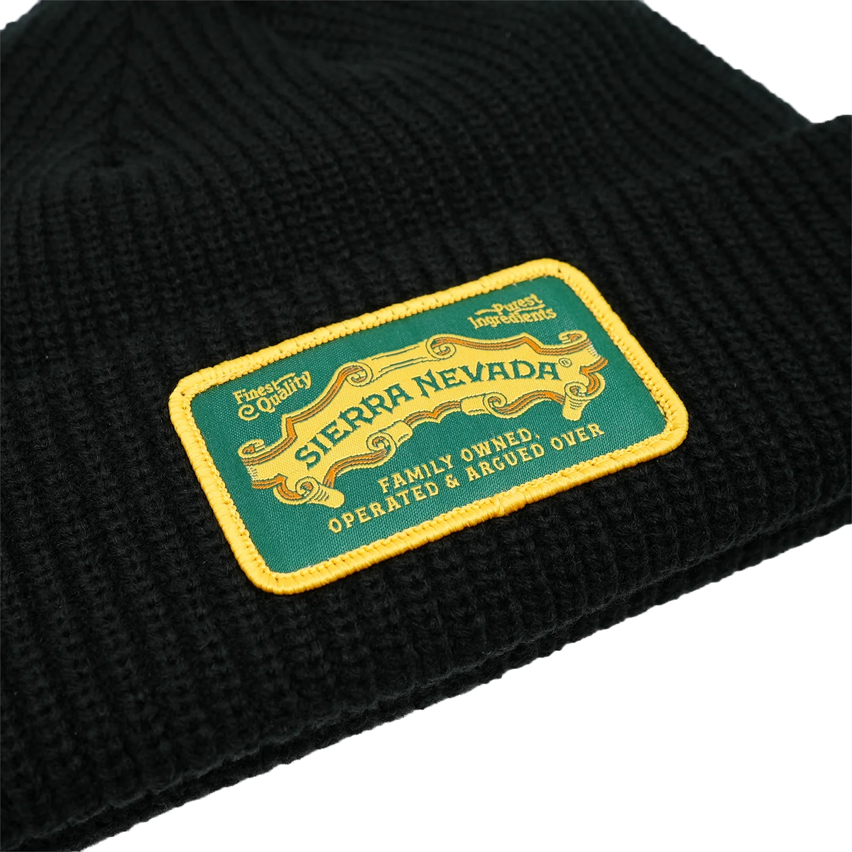 Close-up of a black ribbed knit beanie with a rectangular embroidered patch reading 'Sierra Nevada' in yellow on a green background, with additional text 'Family Owned Operated & Argued Over'