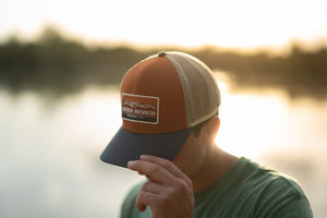 Thumbnail of A man wears the Sierra Nevada Rusty Mountain Gradient Trucker Hat while walking along the banks of a river.