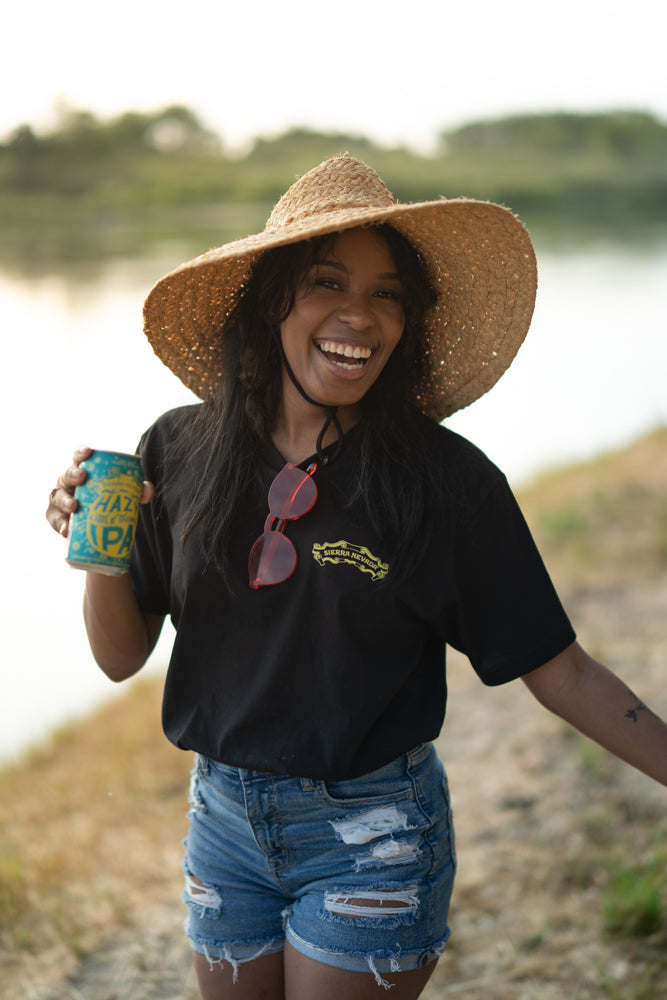 Sierra Nevada Hazy Little Thing black t-shirt, worn by a woman enjoying a Hazy Little Thing IPA on the side of a river.