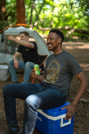 Thumbnail of Sierra Nevada Brewing Co. Chico Facade t-shirt worn by a man sitting on a cooler in a campsite while enjoying a Pale Ale.