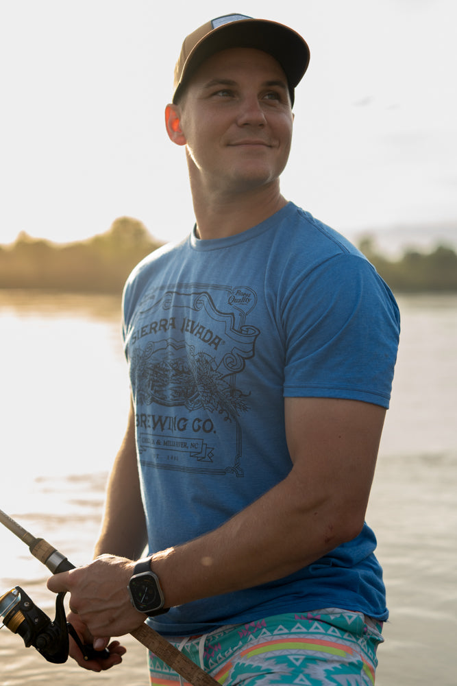 A man wears the Sierra Nevada Brewing Co. Handcrafted T-Shirt while fishing along the riverbank.