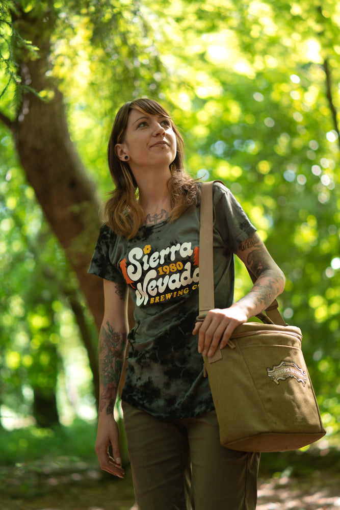 A woman wears the Sierra Nevada Brewing Co. Acid Washed Retro T-Shirt while carrying a cooler and stopping to take in the view in a wooded campsite.