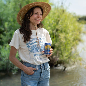 Thumbnail of A woman wears the Sierra Nevada Brewing Co. Handcrafted T-Shirt while enjoying a Summerfest beer at the edge of a river.