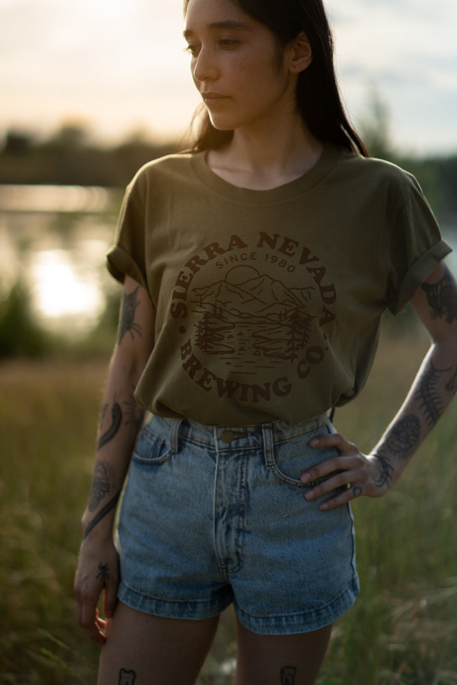 A woman wears the Sierra Nevada Brewing Co. Mountain Circle T-Shirt while standing near a peaceful river.