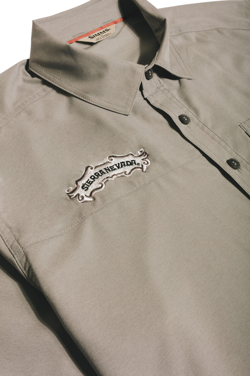 A close-up of a light gray collared button-up shirt with a white embroidered 'Sierra Nevada' logo patch on the chest, set against a dark background.