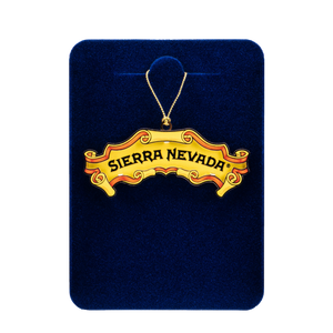 Thumbnail of A gold-tone 'Sierra Nevada' banner ornament necklace displayed on a dark blue velvet backing card against a white background.