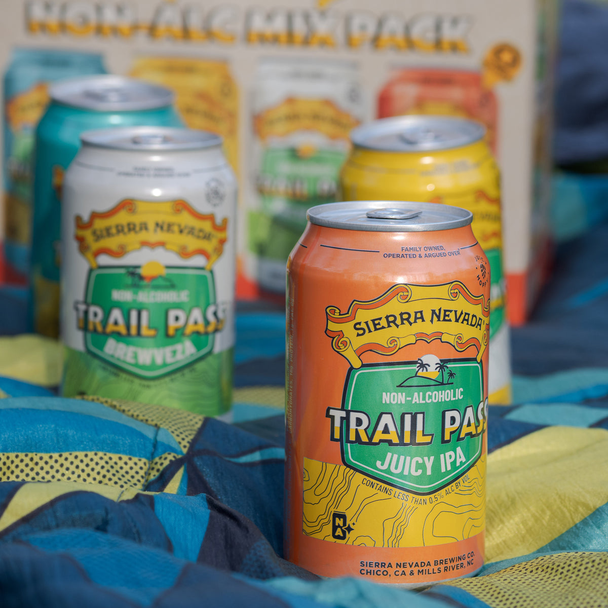 Cans from the Sierra Nevada Trail Pass Variety Pack sitting on a picnic blanket in front of the packaging